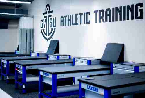 Padded tables are lined up in a room. GVSU Athletic Training is printed on the wall with an anchor.